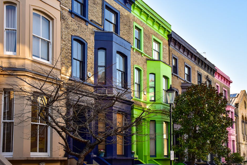 Image of a row of colorful Victorian-style terraced houses on a city street, featuring facades painted in shades of beige, blue, green, and pink. The buildings have large sash windows, some with white frames, and decorative cornices along the rooflines. Leafless trees with sprawling branches are visible in front of the buildings, partially obscuring the lower sections. A lamppost stands near the green building, and the clear sky in the background suggests daytime. The scene captures an urban residential area, which may be part of a home relocation or furniture transport scenario managed by Clapham Man and Van, supporting their removals services for flats near Clapham Common.