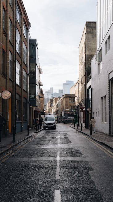 A narrow urban street in Clapham, lined with multi-storey buildings featuring large windows and a mix of brick and modern facades. The street surface appears wet, reflecting the overcast sky above. On the left side, there are shops and a few pedestrians, while on the right side, there are additional buildings with minimal signage. A white van is parked near the middle of the street, and a few bollards line the pavement. In the background, more buildings and a cloudy sky are visible, suggesting a typical city environment suitable for house removals and furniture transport. This scene represents the logistical environment where Clapham Man and Van might assist with packing and loading for house removals, particularly during urban home relocations and moving services in the SW4 area.