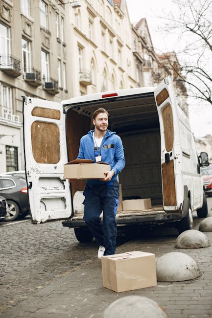 A man wearing a blue work jacket and dark trousers is carrying a medium-sized cardboard box while standing behind the open rear doors of a white moving van parked on a cobblestone street in Clapham. The van's interior is visible, revealing more packing boxes and moving equipment. On the ground nearby, there are additional cardboard boxes and a large round stone. Residential buildings with classic architectural features, such as balconies and window air conditioning units, line the street in the background. The scene captures the process of home relocation, with the man actively engaged in loading or unloading furniture and packing materials as part of a moving service, consistent with professional removals carried out by Clapham Man and Van.