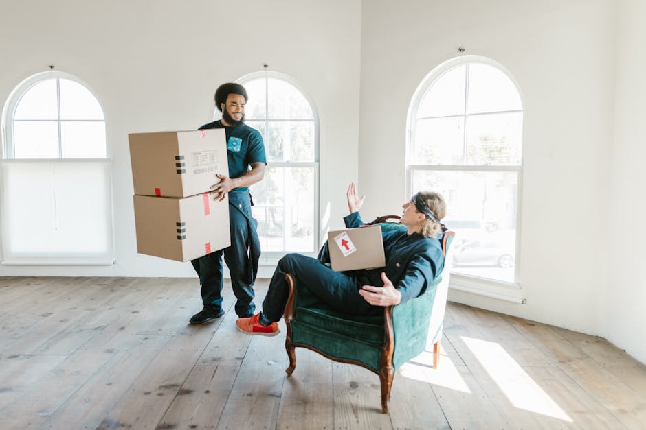 A professional removal team inside a bright, spacious room with large, arched windows allowing natural light to fill the space. One mover, dressed in dark work attire, is carrying two cardboard boxes stacked on top of each other, secured with black and red packing tape. The boxes have red arrow stickers indicating the correct orientation for handling. The other team member, wearing glasses and a blue jacket, is seated comfortably in an upholstered armchair with wooden arms and legs, holding a box on her lap with one arm raised in a gesture of greeting or reassurance. The room has light wooden flooring and minimal furnishings, suggesting a home ready for relocation or packing as part of a home moving or furniture transport process. Clapham Man and Van’s services are implied through the context of packing, load handling, and interior space management, aligned with same-day Clapham removals for emergency flat moves.