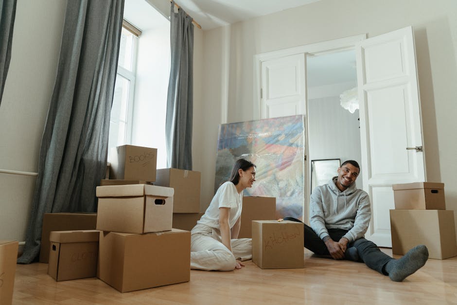 In a brightly lit room with a large window draped with blue curtains, two individuals are sitting on the wooden floor surrounded by packed cardboard boxes, some labelled with words such as 'clothes' and 'books'. An unassembled painting or large artwork leans against the wall, and a small mirror is also visible. The woman, dressed in casual white clothing, and the man, wearing a grey hoodie and dark pants, appear to be in a relaxed moment during a home relocation process. The open white double doors lead into another room, suggesting they are in the midst of packing and preparing for furniture transport or moving tasks. The scene captures elements typical of packing and moving, including cardboard boxes in various sizes, packing materials, and an organized environment for moving house, which Clapham Man and Van offers as part of their same-day house removals for emergency flat moves.