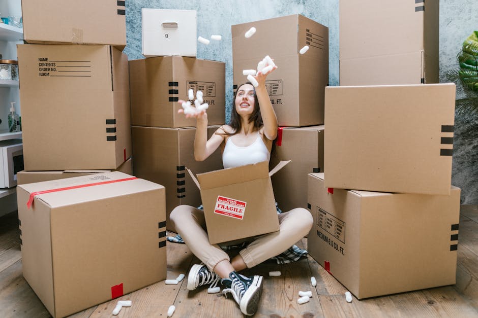 A professional removal team inside a bright, spacious room with large, arched windows allowing natural light to fill the space. One mover, dressed in dark work attire, is carrying two cardboard boxes stacked on top of each other, secured with black and red packing tape. The boxes have red arrow stickers indicating the correct orientation for handling. The other team member, wearing glasses and a blue jacket, is seated comfortably in an upholstered armchair with wooden arms and legs, holding a box on her lap with one arm raised in a gesture of greeting or reassurance. The room has light wooden flooring and minimal furnishings, suggesting a home ready for relocation or packing as part of a home moving or furniture transport process. Clapham Man and Van’s services are implied through the context of packing, load handling, and interior space management, aligned with same-day Clapham removals for emergency flat moves.